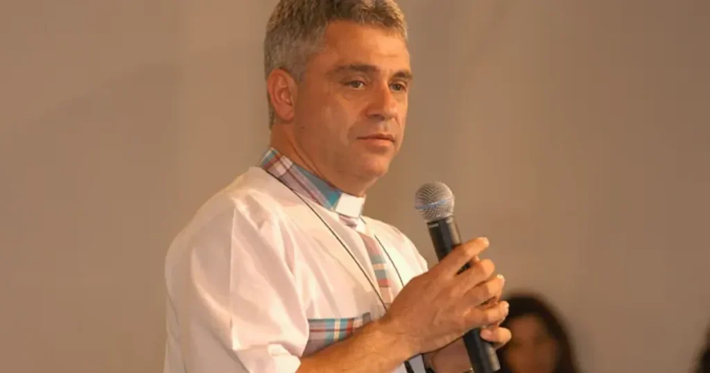 Padre Léo segurando um microfone, com expressão séria e reflexiva, usando batina branca com detalhes xadrez colorido na gola, durante uma pregação.