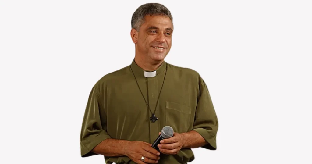 Padre Léo segurando um microfone, sorridente, vestindo batina verde e colar com crucifixo.