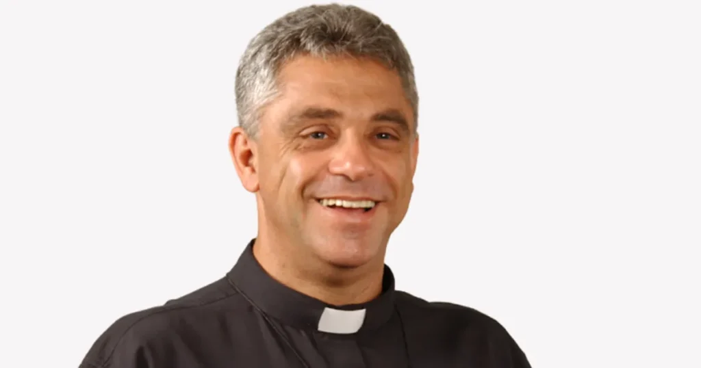 Padre Léo sorrindo, com batina preta clerical e colar com crucifixo, em fundo branco.